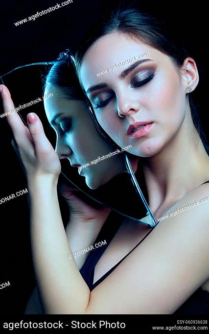 Girl with a shard of the mirror. Female with mirror shard in hand posing on gray background. Face reflection in mirror splinter. Eyes closed