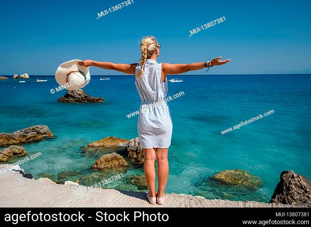 A tourist woman with hat raising her hands against sea at Agios Nikitas village on Lefkas Ionian Island, Greece