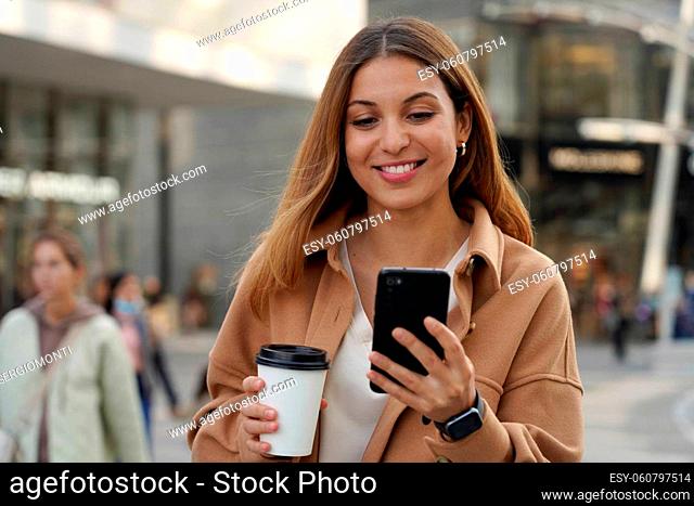 Young multi ethnic business woman walks watching mobile phone with blurred people on modern urban background