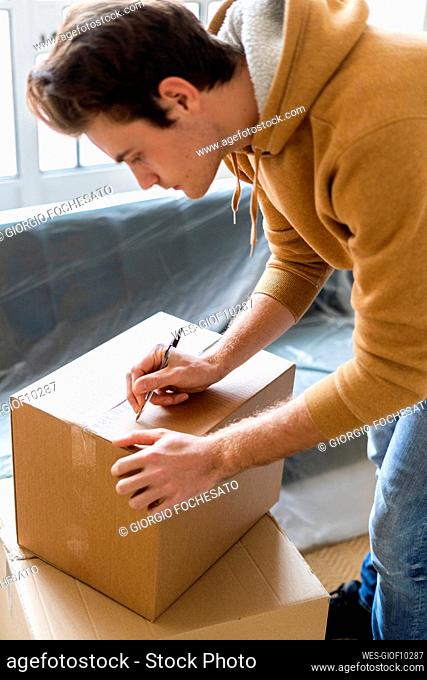 Young man writing on cardboard box while moving into new loft apartment
