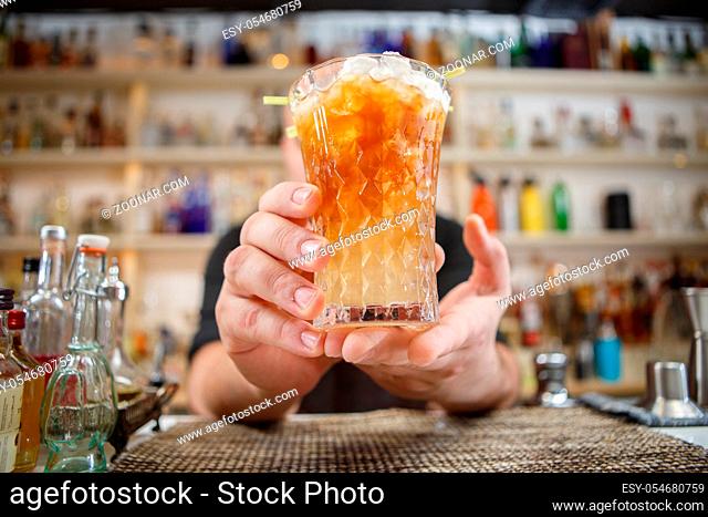 Bartender is standing in pub? holding cocktail and giving glass forward. Focus on beverage
