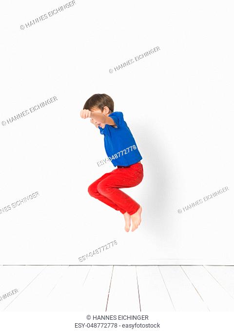 cool, young and cute boy with blue shirt and red trousers is jumping high in the studio in front of white background and white wooden floor