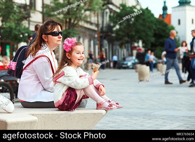 Mother and daughter spending time together, sitting in the center of town, little smiling girl holding and eating ice cream