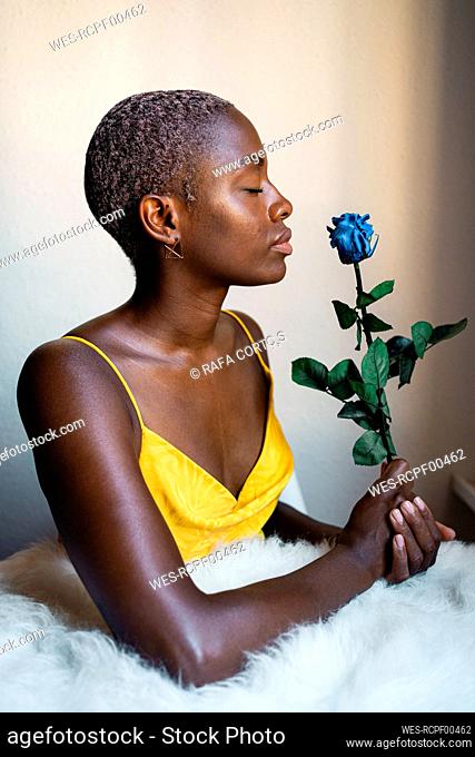 Woman with eyes closed holding blue rose against wall at home