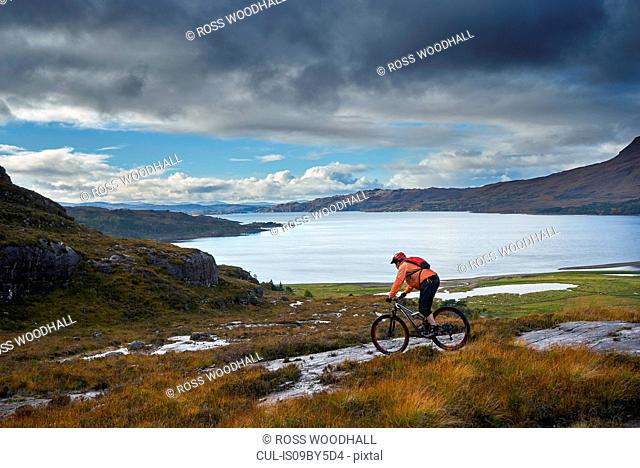 Male mountain biker biking down toward loch in mountain landscape, Achnasheen, Scottish Highlands, Scotland