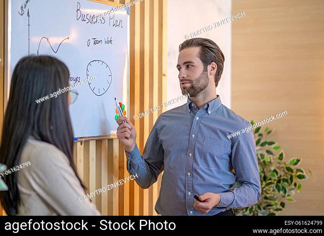 Male office worker pointing with a marker at the circular diagram on the whiteboard to his female colleague