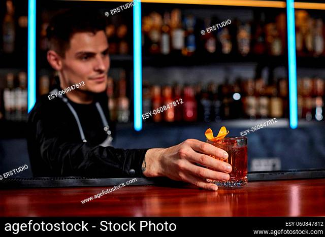 Barman in apron holding out glass with alcoholic cocktail to the client. Bartender is making cocktail