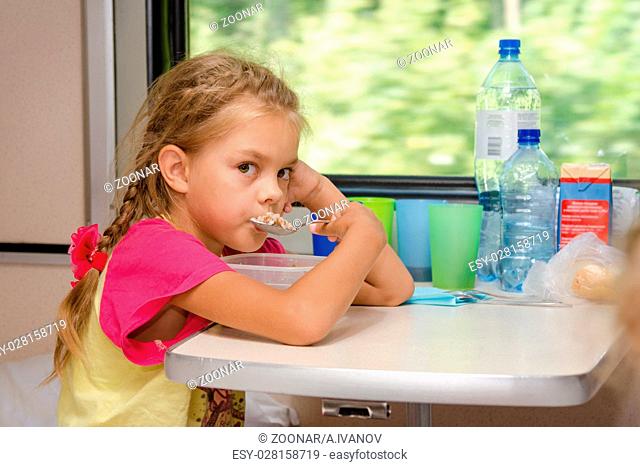 A six-year girl in a train sitting at the table on the lower place in the second-class compartment of the car and eats porridge spoon