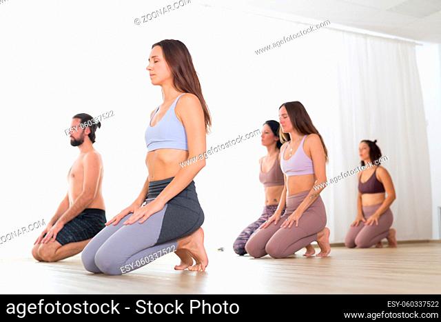 Group of young sporty attractive women in yoga studio, practicing yoga lesson with instructor, kneeling down, stretching and relaxing after workout