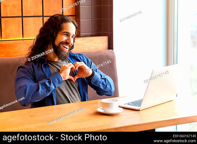 Portrait of romantik handsome young adult man freelancer in casual style sitting in cafe and talking with his friend in laptop, showing heart sign with fingers