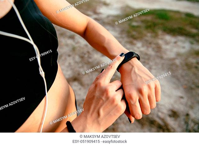 Young female checking the time on her watches after running, outdoors, listening to music in headphones. Workout concept. Close-up view