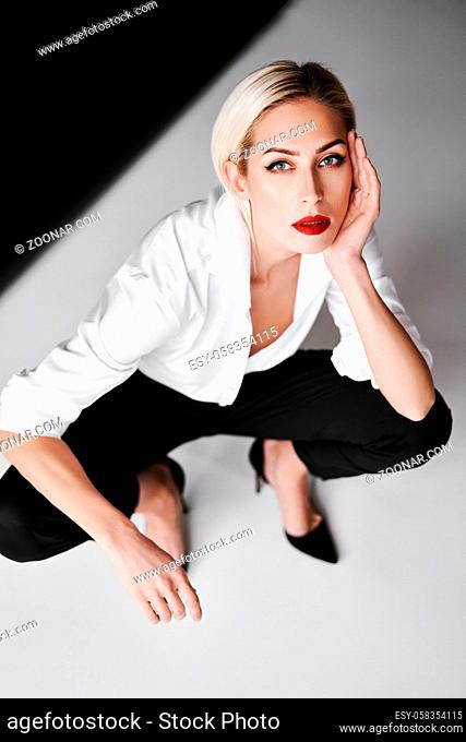 Top view portrait of daring trendy woman posing on white studio background sitting on floor. Gorgeous stylish blonde female wearing white shirt