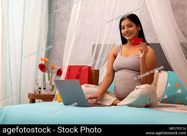Smiling pregnant woman shopping online on laptop using credit card while sitting on bed at home