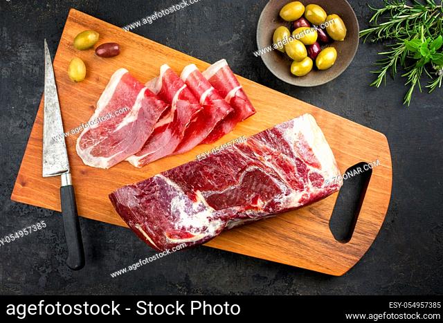 Traditional dry cured and smoked ham with herbs and olives offered as top view on a modern design cutting board