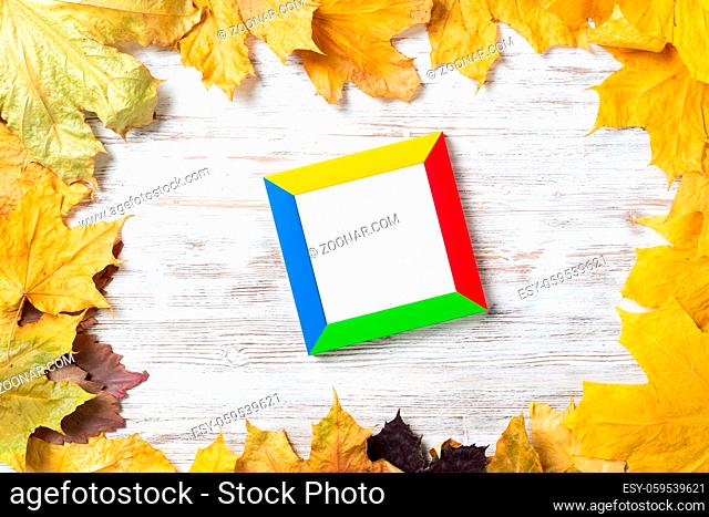 Blank rectangular photo frame lies on vintage wooden desk with bright autumn foliage. Flat lay with autumn leaves on white wooden surface