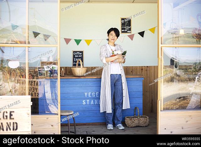 Japanese woman standing in a farm shop, holding vegetables, smiling at camera