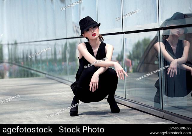 Stylish asian girl in a black suit and hat, posing with a glass wall