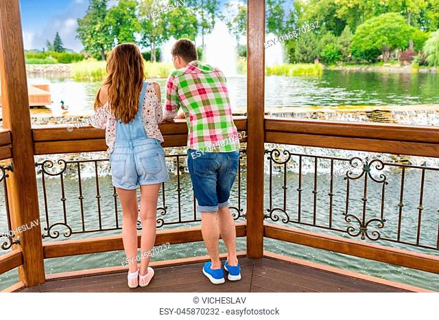 Back view of young male and female leaning on railing of the wooden gazebo standing in front of human-made waterfall in a park with pond