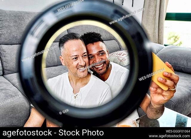 Smiling couple taking selfie seen through ring light in living room
