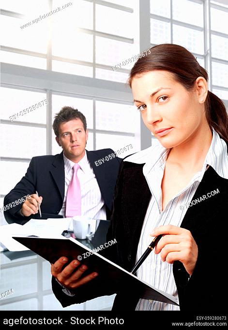Young secretary writes notes into notepad while a businessman awaits in the background