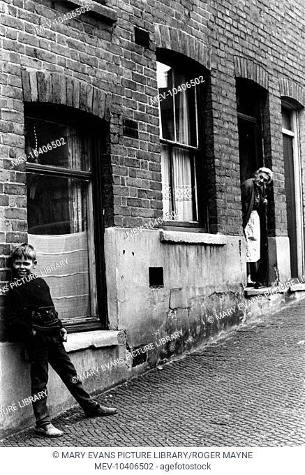 Scene in a slum street in the St Ann's area of Nottingham. A young boy leans against the wall while an old lady looks out of her door at him