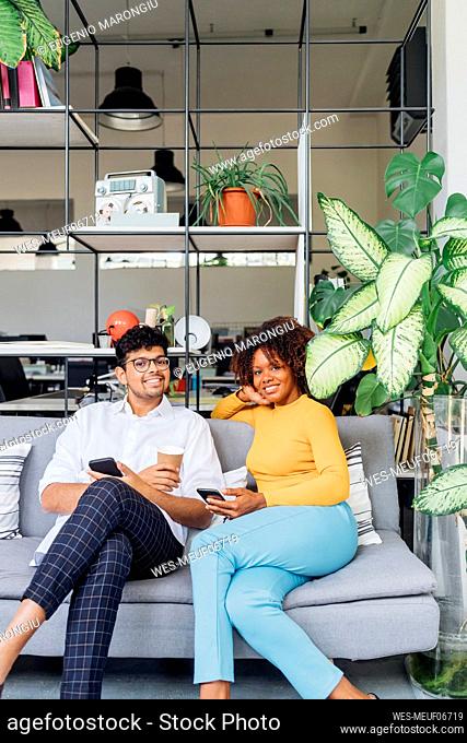 Smiling colleagues with mobile phones on sofa at workplace
