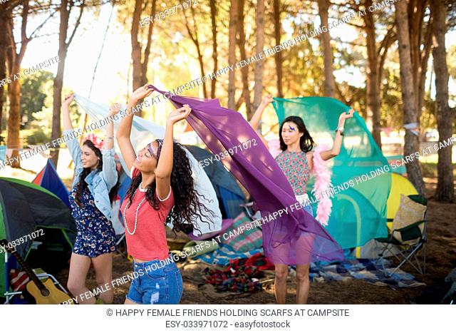 Happy female friends holding scarfs while standing on field at campsite