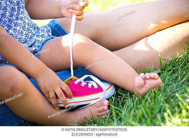 Caucasian Little toddler girl with blue free playing on her mom's lap on the grass and tiying her mini converse pink shoe