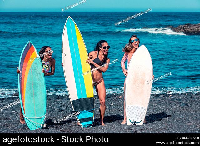 Group of laughing diverse women with surfing boards standing together on seashore in bright sunlight