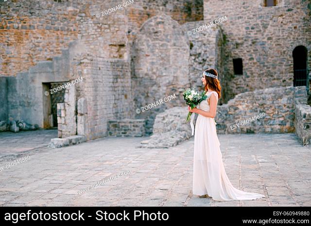 The bride stands on the square near the castle walls in the old town of Budva and holds a bouquet in her hands . High quality photo