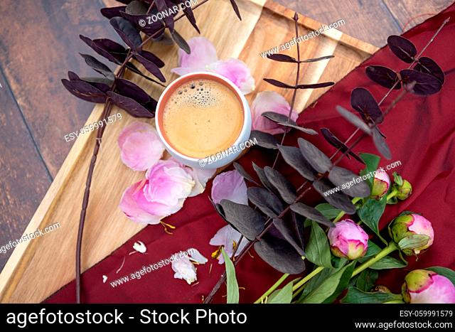 A white Cup of black coffee stands on a wooden background next to pink peonies and other garden flowers and leaves, The concept of village life, Cottagecore