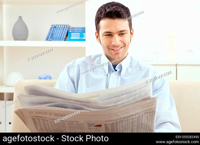 Happy young man sitting on couch reading newspaper at home, smiling