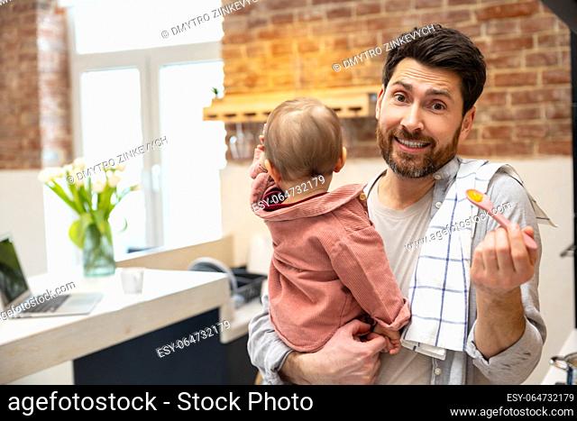 Father with little baby girl in arms in kitchenn feeding daughter with fruit or vegetable puree, complementary feeding of child