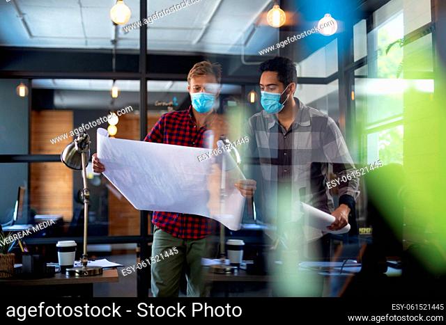 Diverse male colleagues wearing masks in disucssion holding architectural plans in office
