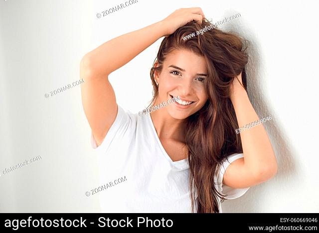 Young smiling playful blond woman holding her hair while leaning against white wall