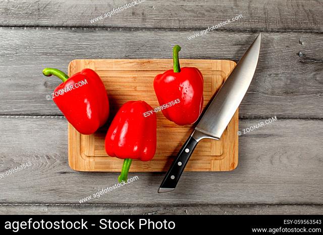 Table top view - three bright red bell peppers, water drops, with chefs knife on chopping board, with rustic gray wood desk under