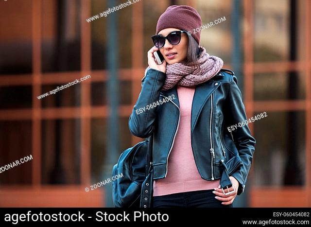 Young fashion woman in black leather jacket using cell phone in city street Stylish female model wearing sunglasses beanie and scarf outdoor