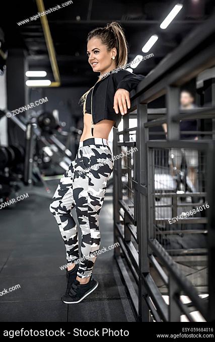 Beautiful sportive girl leans on the railing in the gym. She looks to the side with a smile. Woman wears light pants with prints
