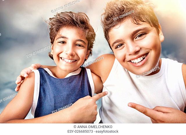 Two cute cheerful boys hugging and showing on each other by finger, portrait over cloudy sky background, happy children having fun at summertime
