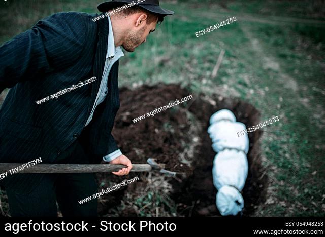 Maniac with a shovel buries victim wrapped in a canvas into a grave, serial murderer concept, crime horror