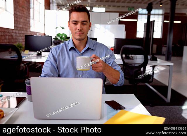 Caucasian businessman sitting at desk using laptop and drinking cup of coffee