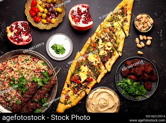 Various Turkish dishes: pide pizza, meat kebab with tabbouleh salad, falafel, hummus, olives, pistachios and Middle Eastern meze on black concrete table top...