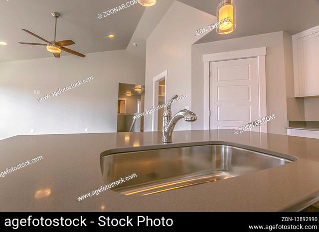 Gleaming stainless steel sink and faucet inside the kitchen of a new home. Warm celiling lights and a ceiling fan can also be seen inside the house
