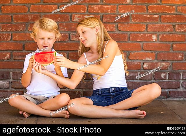 Cute young cuacasian boy and girl eating watermelon against brick wall