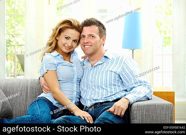 Portrait of happy couple sitting on sofa embracing, looking at camera and smiling