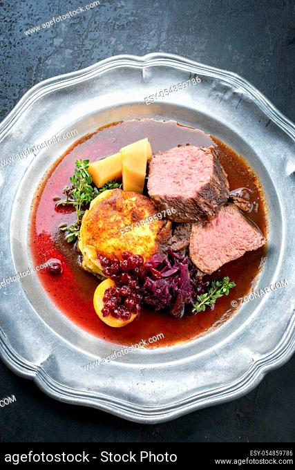 Traditional saddle of venison with fried mashed potatoes and red cabbage in game red wine sauce as top view on a pewter plate