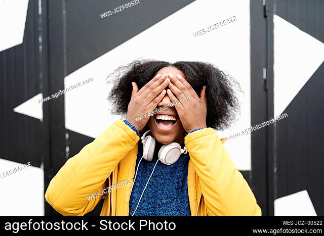 Happy woman covering eyes with hands against wall