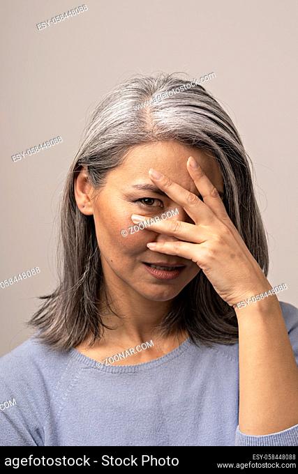 Middle-Aged Asian Woman Covers Her Face With Hand And Looks Displeased. Dissatisfied Grey-Haired Woman Who Covers Her Left Side Of Face. Portrait