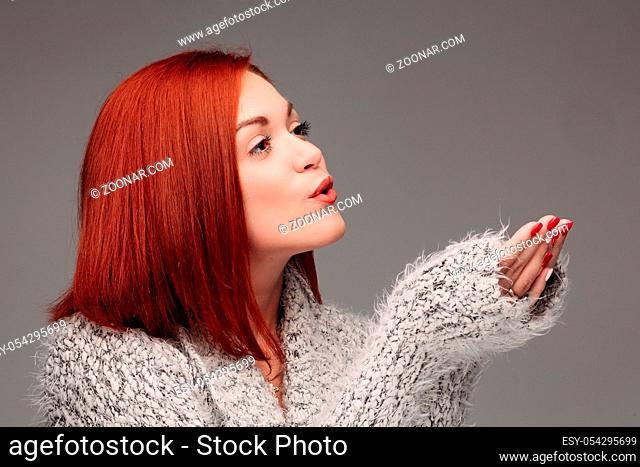 Beautiful young girl with red hair and red nails holding her hands together and blowing on white lightening ball. Pretty woman in grey sweater catching star and...