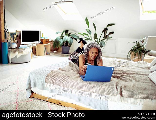 Senior woman with credit card paying bills at laptop on bed
